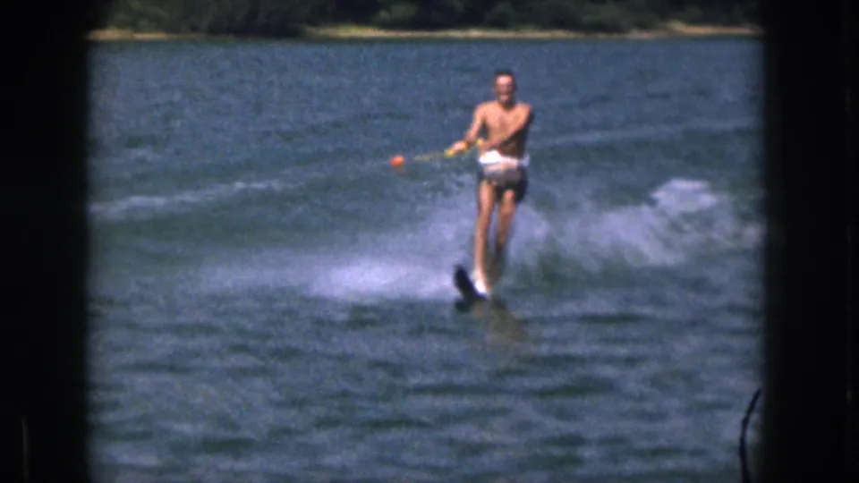 In 1961 Michigan home-movie footage, a shirtless adult man in swim trunks rides water skis across a lake while holding a tow rope with an orange float nearby.