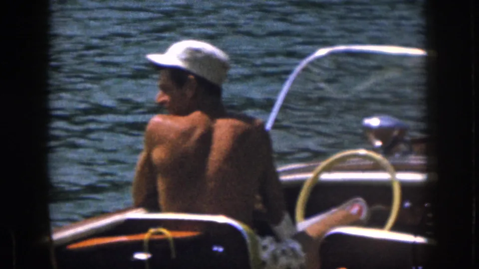 From the boat, a shirtless adult man in a white cap sits near the steering wheel and looks back over the lake during the water-skiing outing.