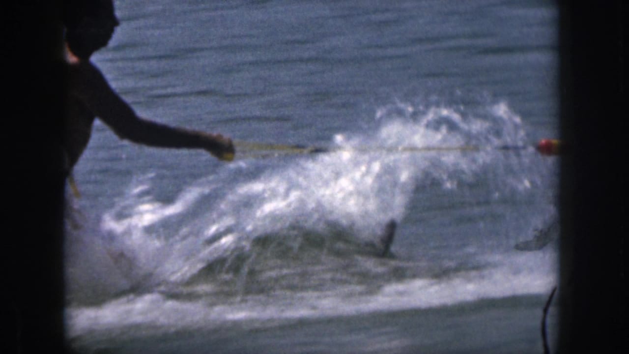 1961 Michigan 8mm frame of an adult water skier at the edge of view, pulled across lake water with a large white spray.