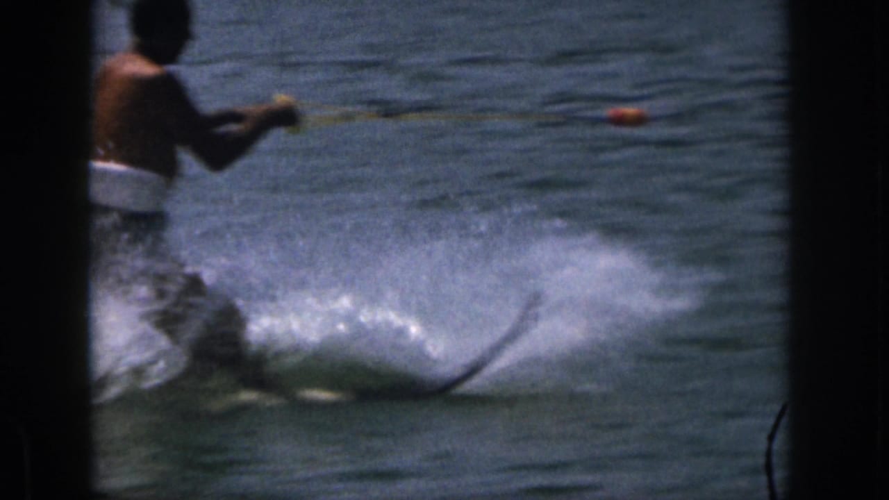 A shirtless water skier in dark trunks holds a tow rope while skimming over lake water.