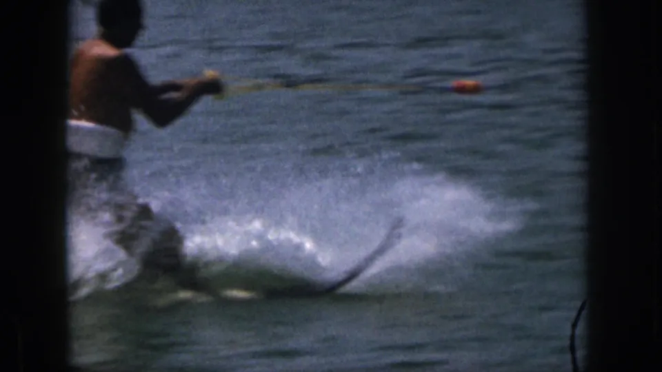 1961 Michigan home-movie frame showing a shirtless adult water skier in dark trunks and a white flotation belt on the lake.