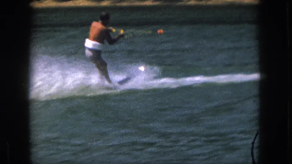 1961 Michigan home-movie frame of an adult man water skiing across open lake water with tow rope, spray, and orange buoy visible.