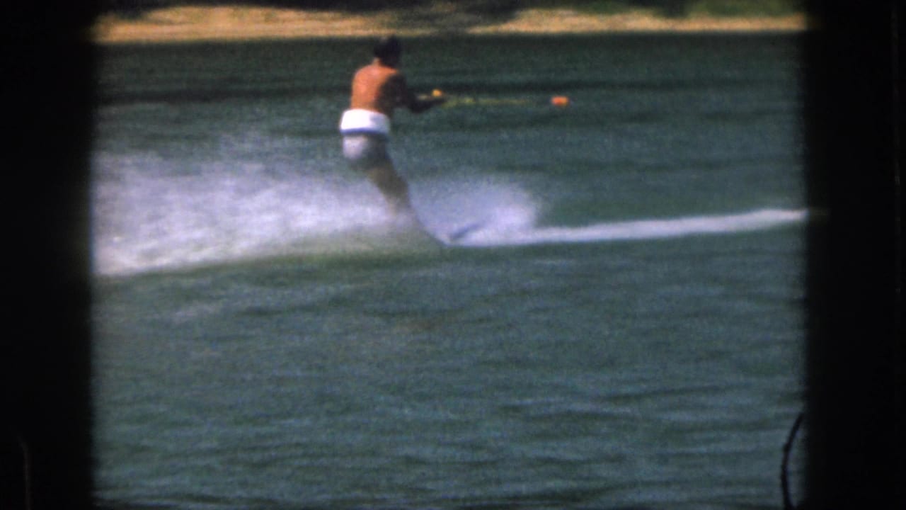 1961 Michigan 8mm frame of a shirtless man in white trunks water skiing across a lake with spray trailing behind him.
