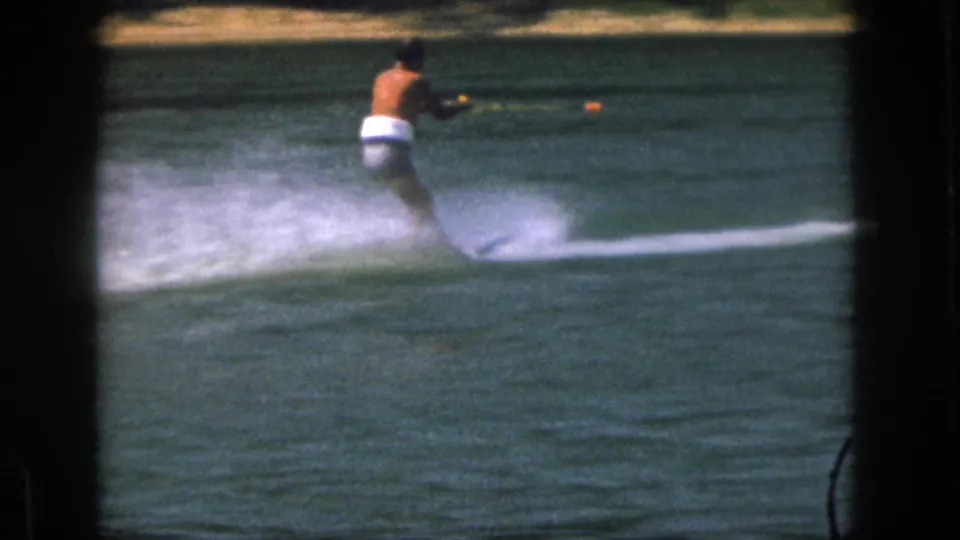 1961 Michigan 8mm frame of a shirtless man in white trunks water skiing across a lake with spray trailing behind him.
