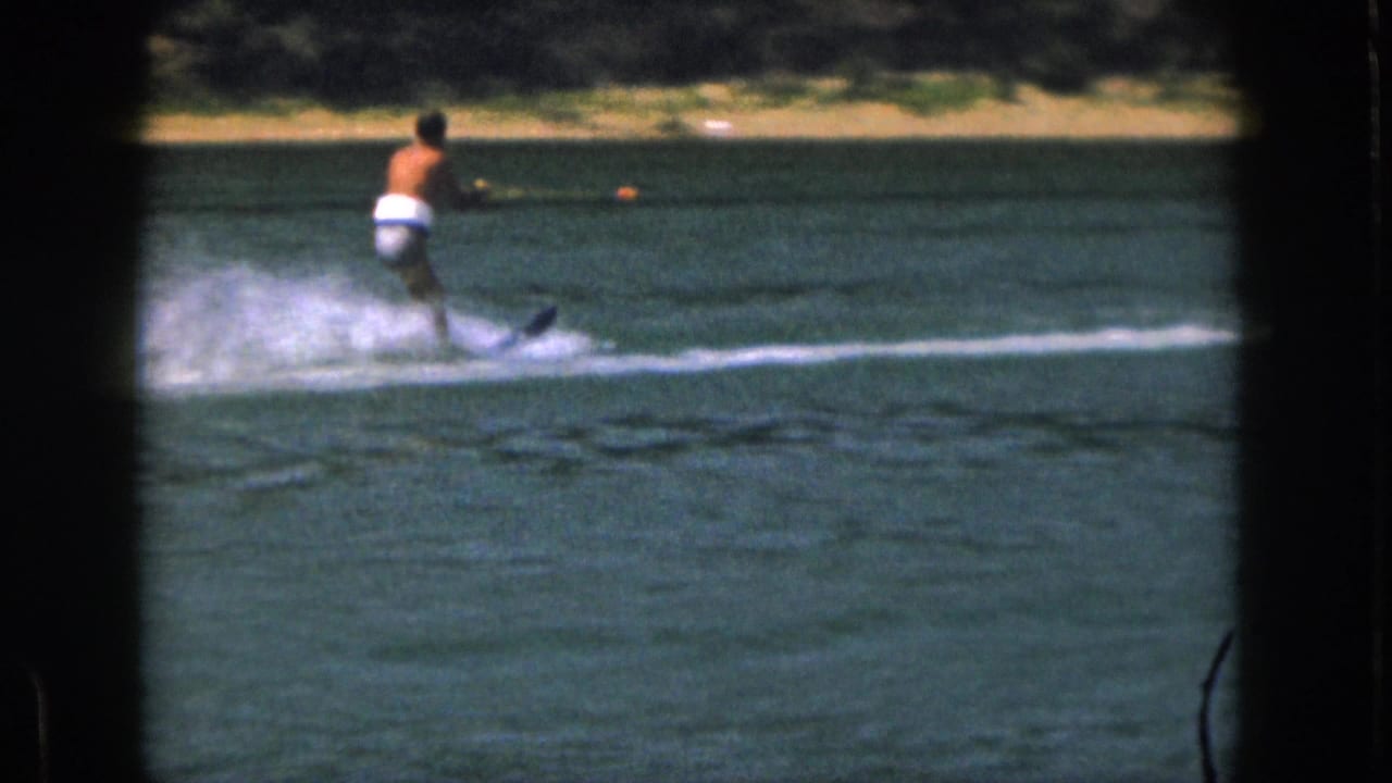1961 Michigan home-movie frame showing a distant water skier holding a tow rope as his ski cuts through the lake.