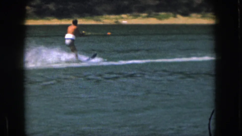 1961 Michigan home-movie frame showing a distant water skier holding a tow rope as his ski cuts through the lake.