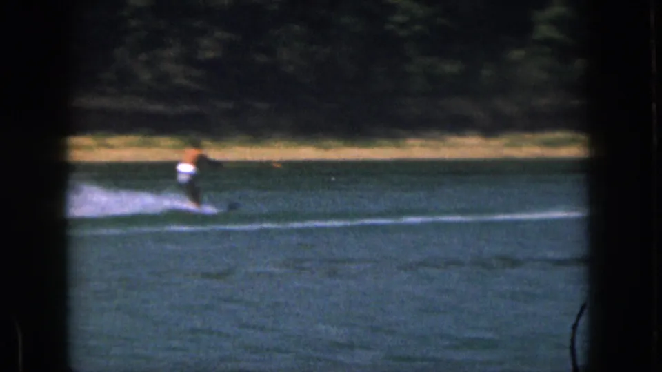 1961 Michigan home-movie frame with a shirtless water skier crossing a lake, framed by dark 8mm borders and a wooded shore.