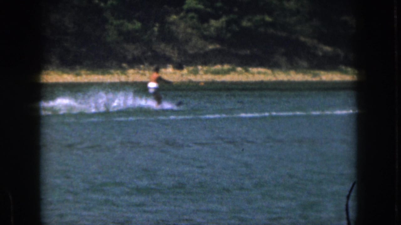 Distant water skier on a Michigan lake in 1961, seen through dark 8mm frame borders with shoreline trees behind.