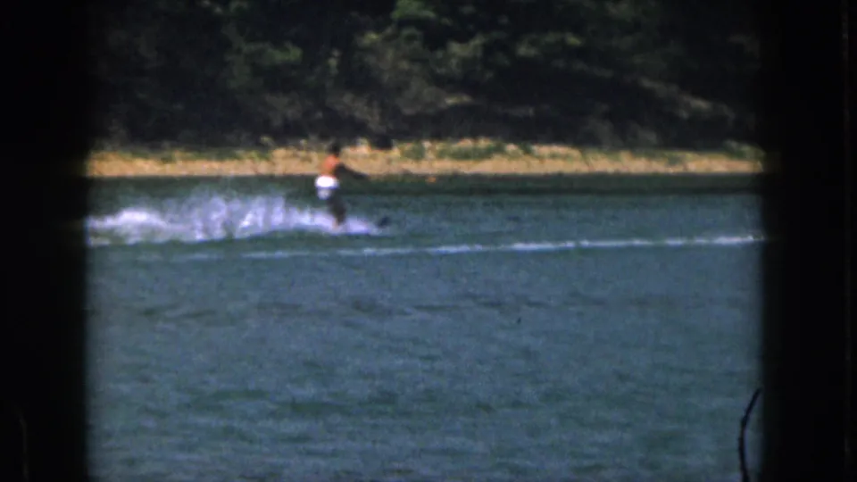 Distant water skier on a Michigan lake in 1961, seen through dark 8mm frame borders with shoreline trees behind.