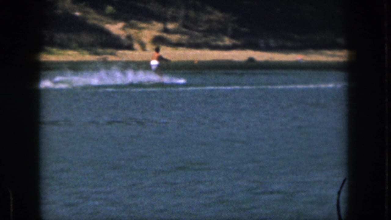 The skier continues across calm Michigan lake water, with a sandy bank and dark wooded shore behind the spray.