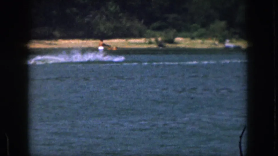 A small distant skier throws a broad spray while crossing the lake, framed by soft 8mm color and a wooded Michigan shoreline.
