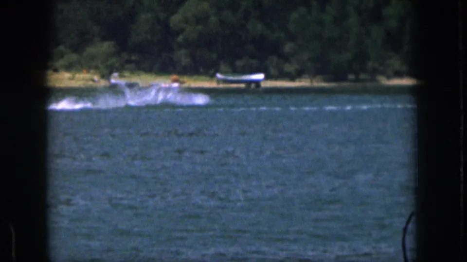 The skier or tow path nears the frame edge as spray and a small distant boat appear along the Michigan lake shoreline.