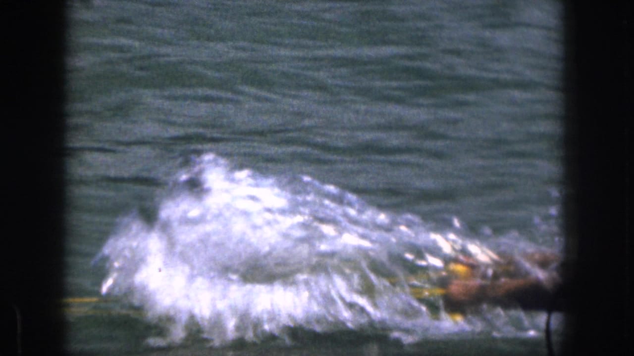 1961 Michigan home-movie frame showing a waterskier low in the water beside heavy spray and a yellow tow rope.