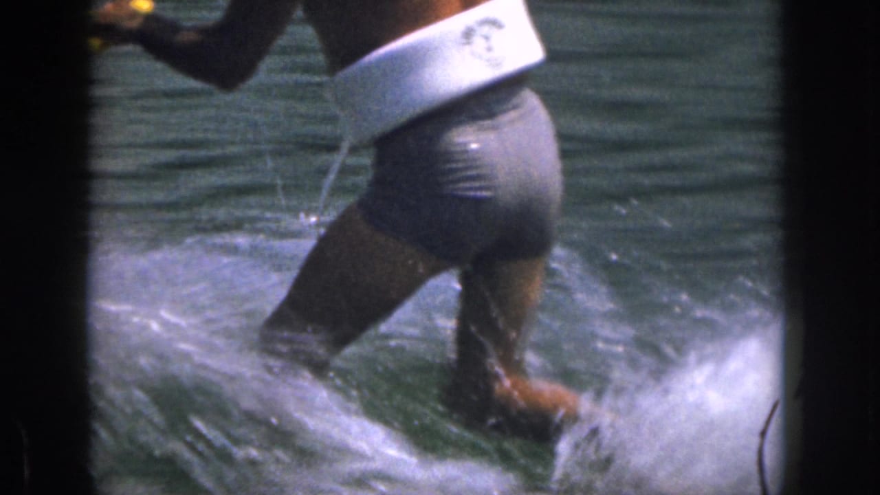 Man in a white life vest and dark trunks waterskiing across a lake behind a tow rope.
