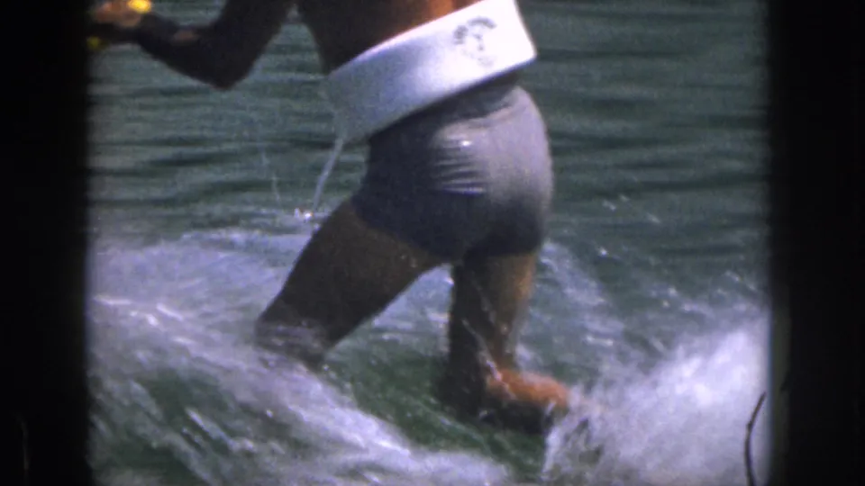 1961 Michigan frame of a man waterskiing across a lake in a white flotation vest and dark swim trunks, with spray behind him.