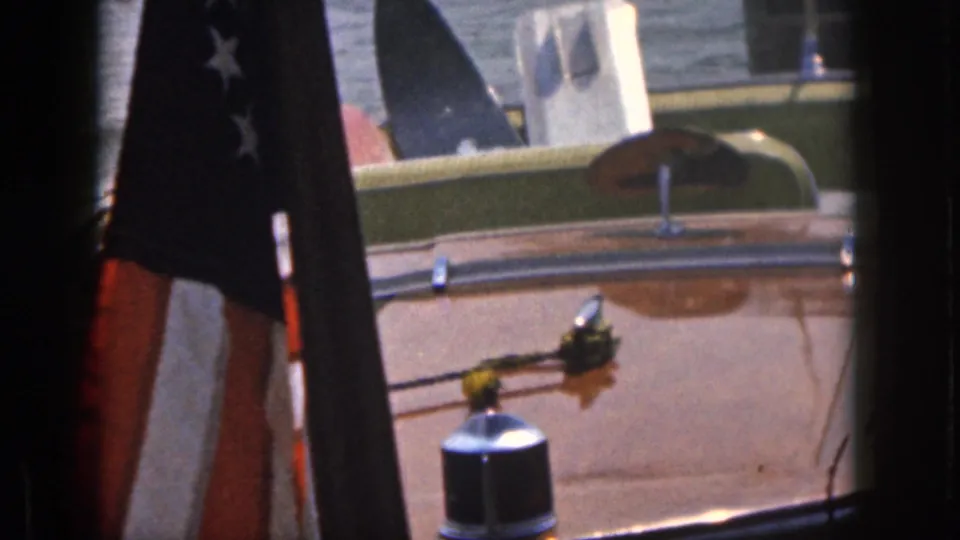 1961 Michigan home-movie frame of a varnished boat deck with an American flag, green interior trim, chrome fittings, and water behind.