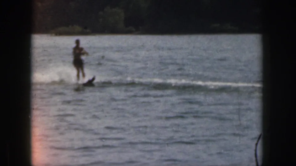 Distant waterskier on a Michigan lake in 1961 home-movie footage, moving across the water with spray and shoreline in the background.