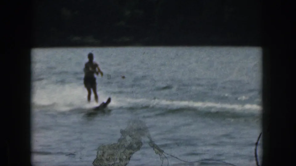 1961 Michigan waterskiing frame showing a distant skier riding across open lake water, with spray, wake, and a dark shoreline beyond.
