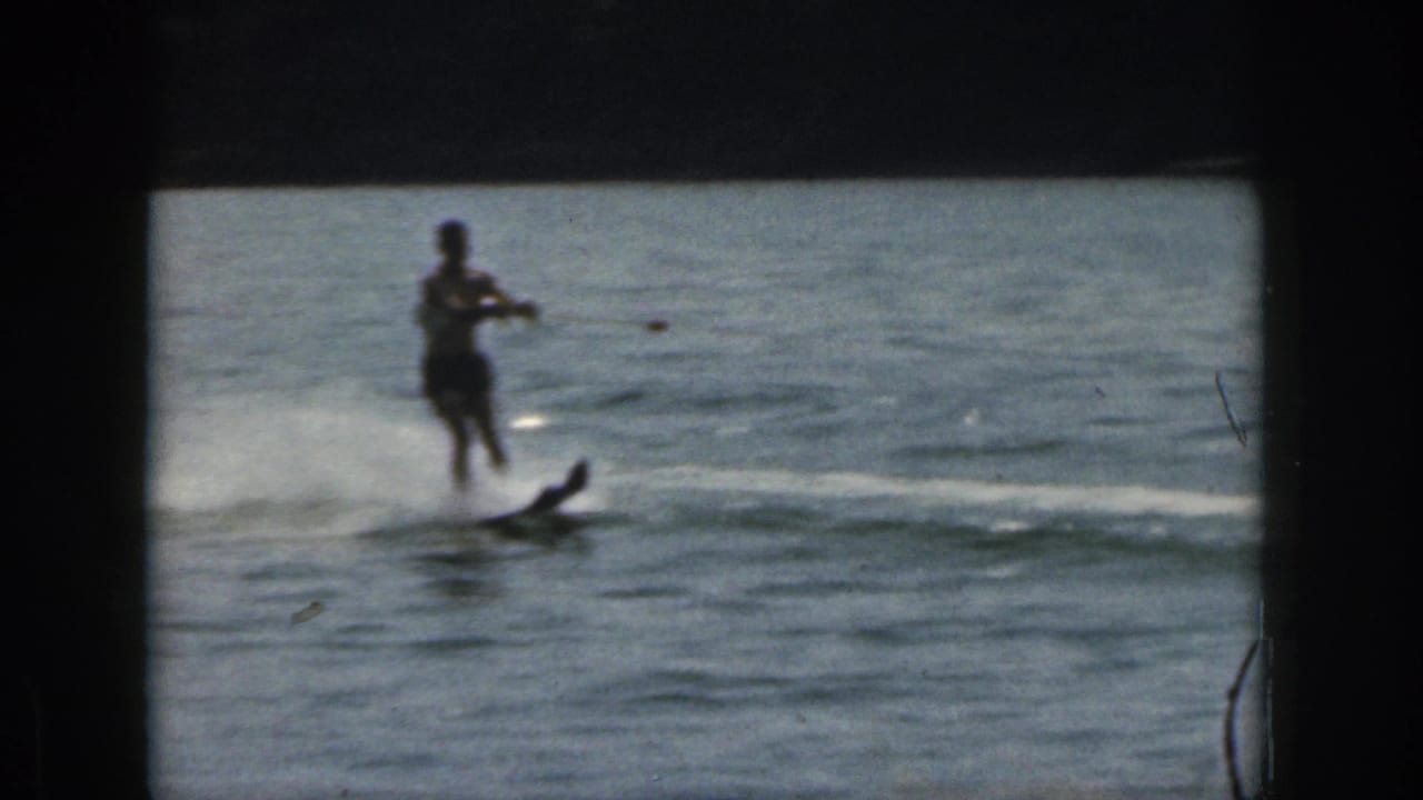 1961 Michigan home-movie frame showing a man water skiing across a lake, with white spray, soft focus, and dark 8mm frame edges.