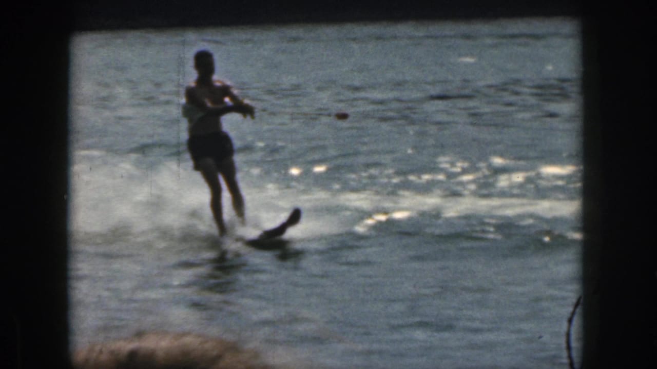 Man in dark swim trunks water skiing on a Michigan lake in 1961, seen through grainy 8mm color footage with bright water reflections.