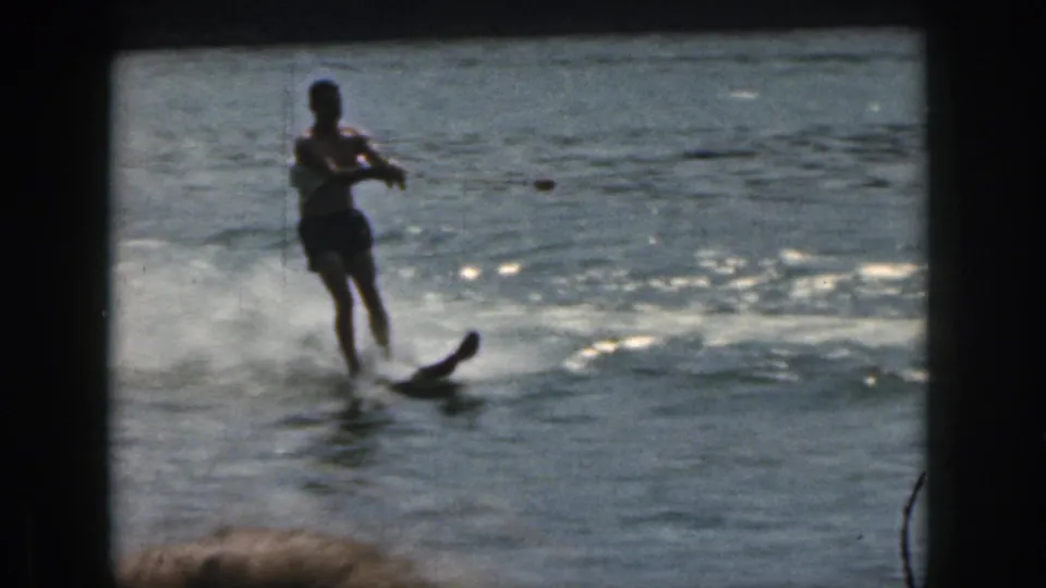 Man in dark swim trunks water skiing on a Michigan lake in 1961, seen through grainy 8mm color footage with bright water reflections.