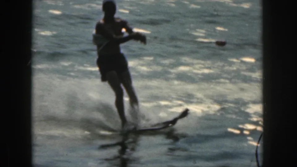 Closer 8mm frame of a man water skiing on a lake in Michigan, 1961, leaning back with spray and reflected light around the ski.
