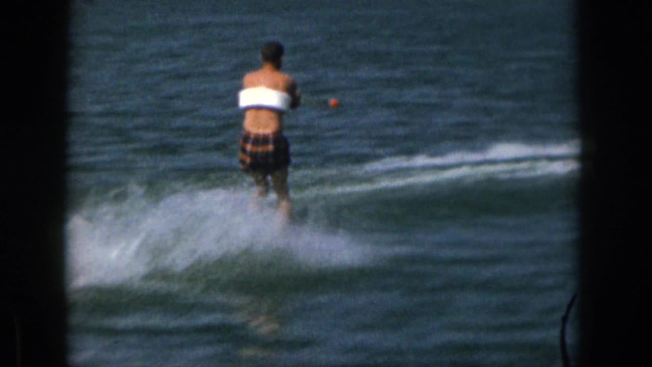 1961 Michigan home-movie frame of a man water skiing away from the camera on a lake, with dark film borders and shoreline in the distance.