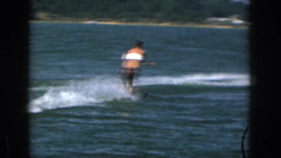 A distant water skier in a white life vest glides across a Michigan lake in 1961, framed by dark 8mm film edges.