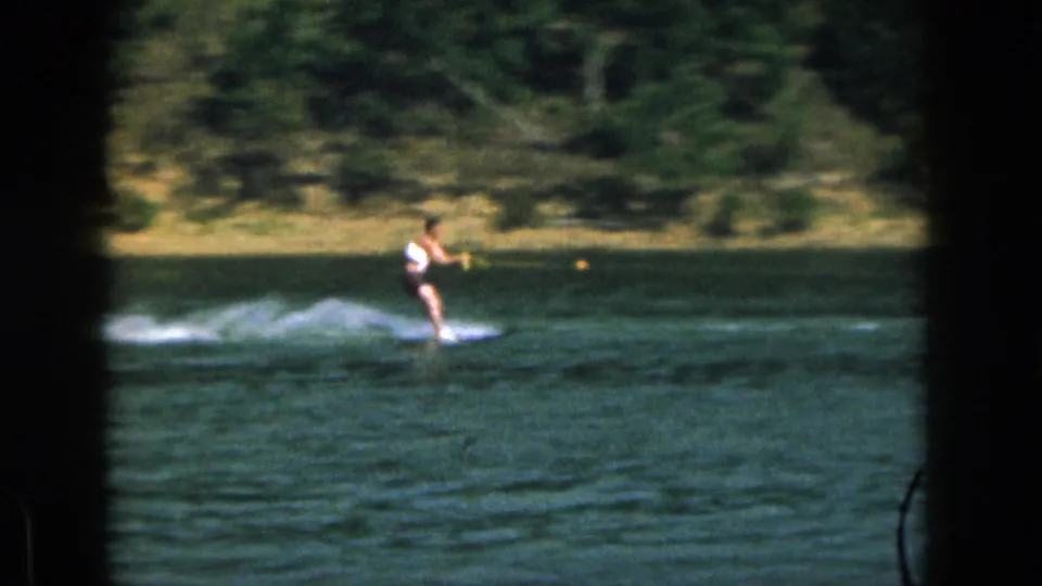 1961 Michigan 8mm frame showing a water skier at mid-distance, with lake spray and a tree-lined shore behind him.