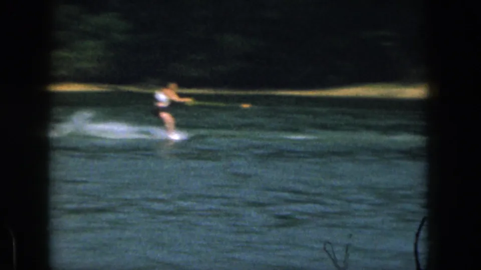 A distant water skier crosses a Michigan lake in faded 8mm color, with shoreline and dark film borders visible.