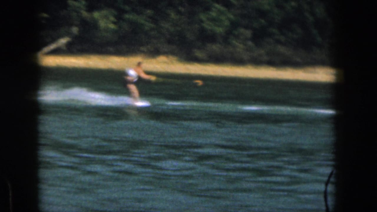 1961 Michigan 8mm frame showing a distant water skier crossing a calm lake with a tow rope and white spray behind him.