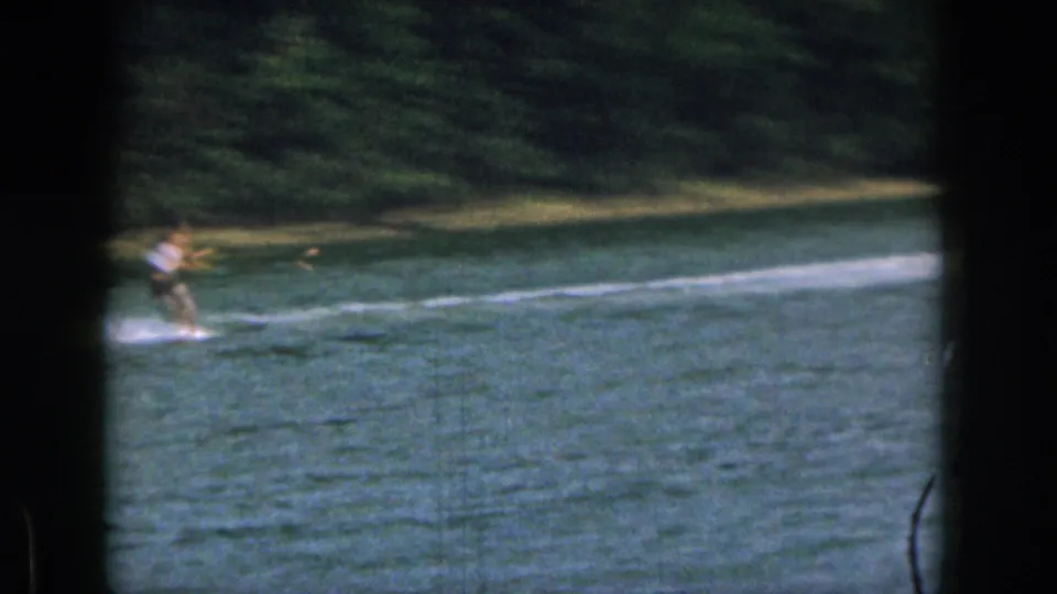 A distant skier in light and dark clothing rides across a Michigan lake, with wake trailing behind and trees along the far bank.