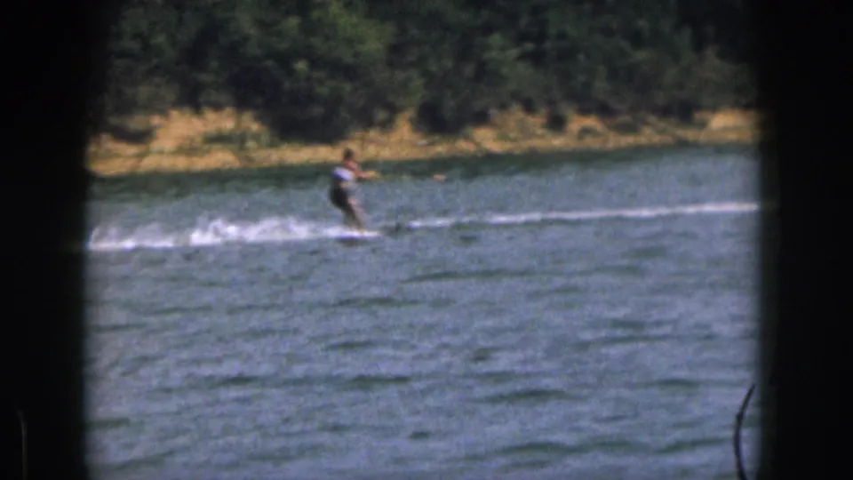 A 1961 home-movie frame shows the skier leaning over the water as spray breaks behind him near the wooded shoreline.