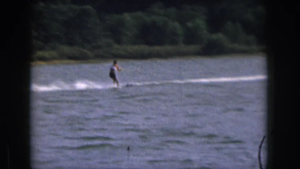 The skier appears farther from camera on the lake, leaving a long white wake against the wooded Michigan shoreline.