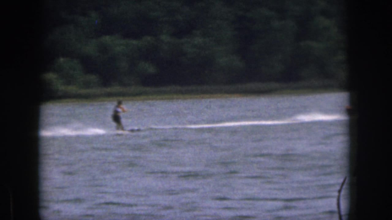 1961 Michigan home-movie frame showing a distant water skier gliding across a calm lake near a tree-lined shore.