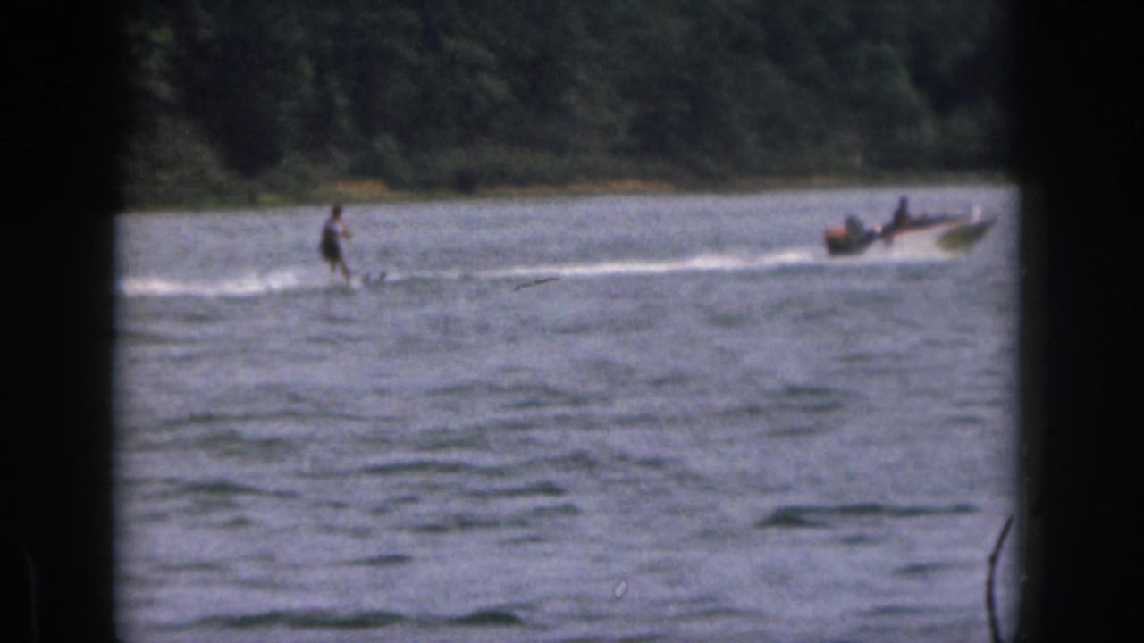 A water skier follows a motorboat across the lake near a wooded shore.