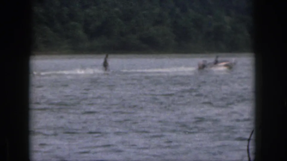 Distant 1961 Michigan home-movie view of a skier and tow boat crossing a wooded lake scene.