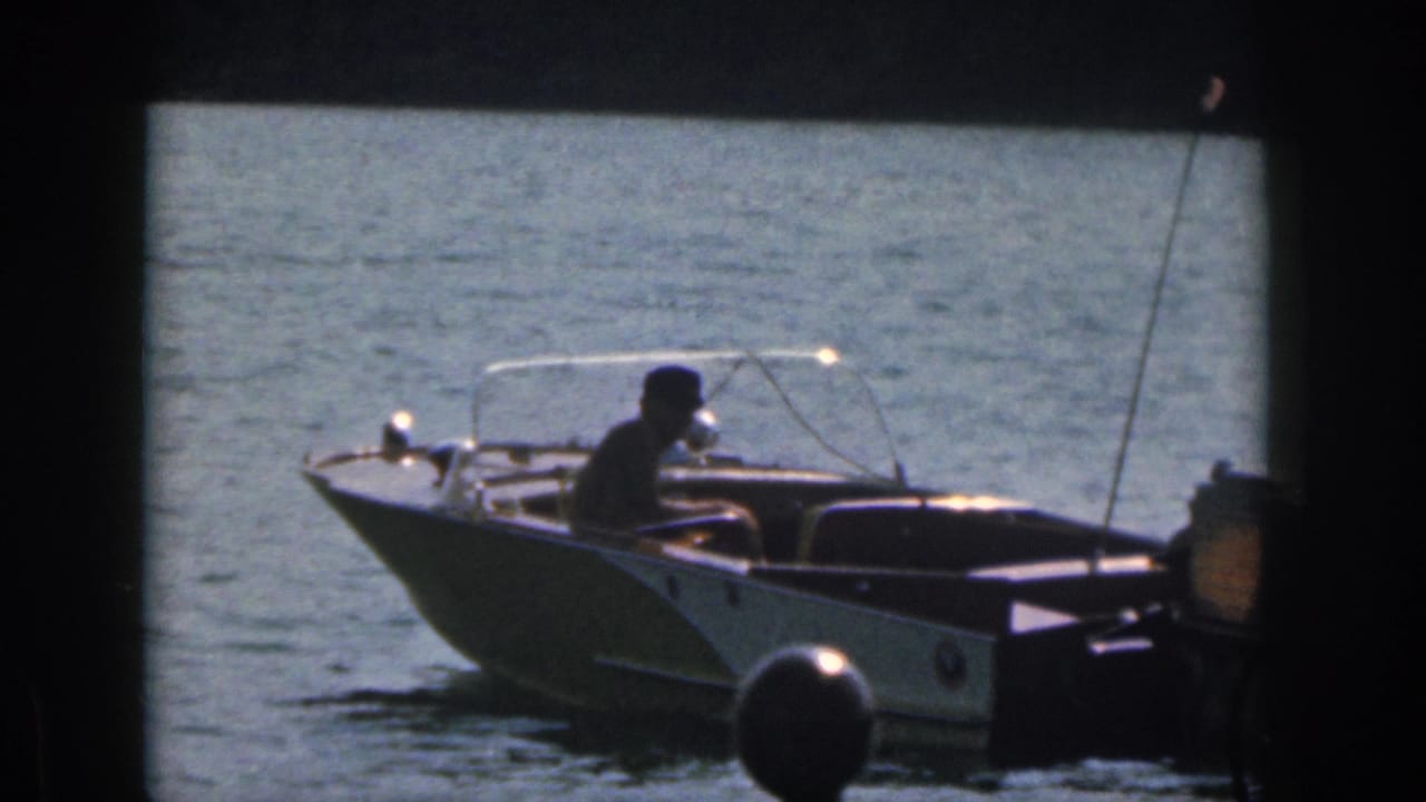 1961 Michigan 8mm frame of a man seated at the helm of a small motorboat moving across blue water.
