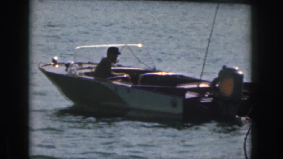The home-movie frame shows a man steering a small runabout, with an outboard motor visible at the stern.