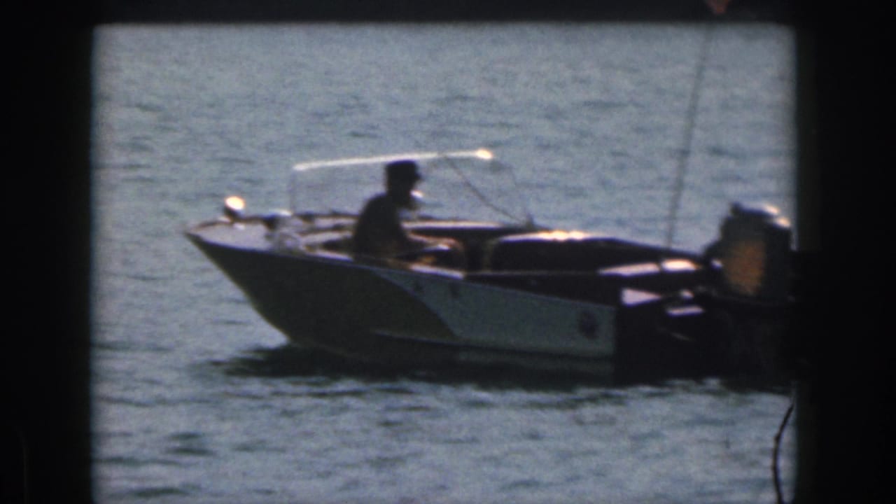 1961 Michigan home-movie frame showing a small open motorboat on the lake with a seated person near the controls.