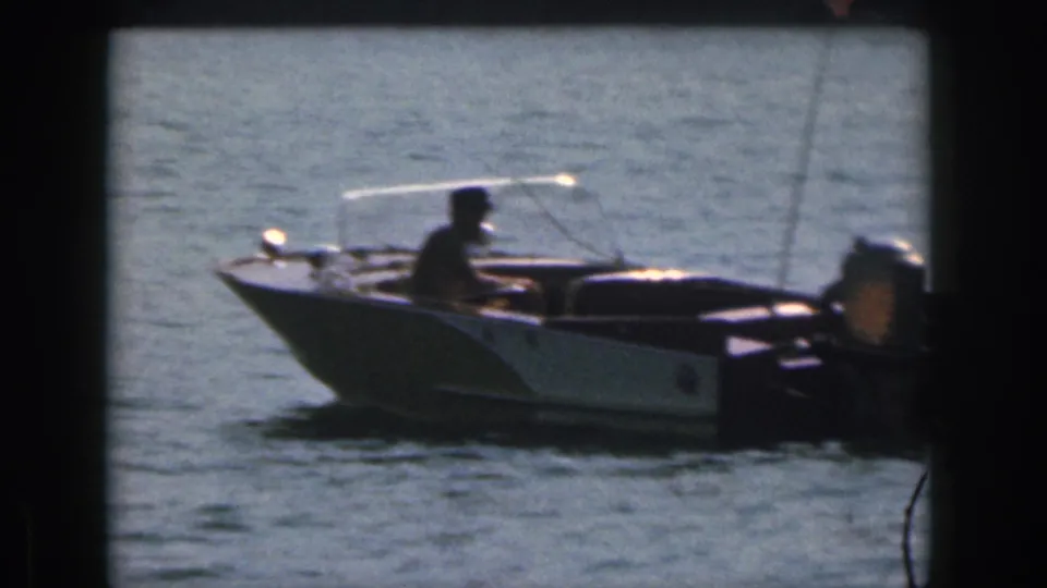 1961 Michigan home-movie frame showing a small open motorboat on the lake with a seated person near the controls.