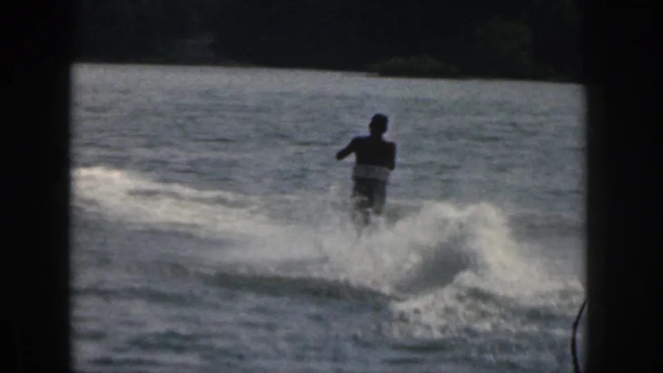 1961 Michigan home-movie frame with a distant silhouetted water skier moving across the lake inside a dark 8mm frame edge.