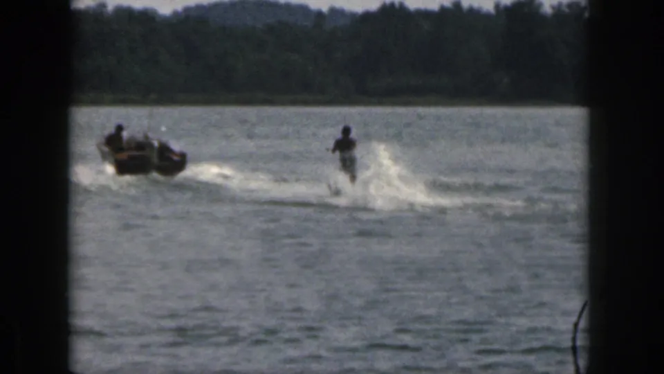 A small boat tows a water skier across a Michigan lake in 1961, with spray and wooded shoreline in the background.