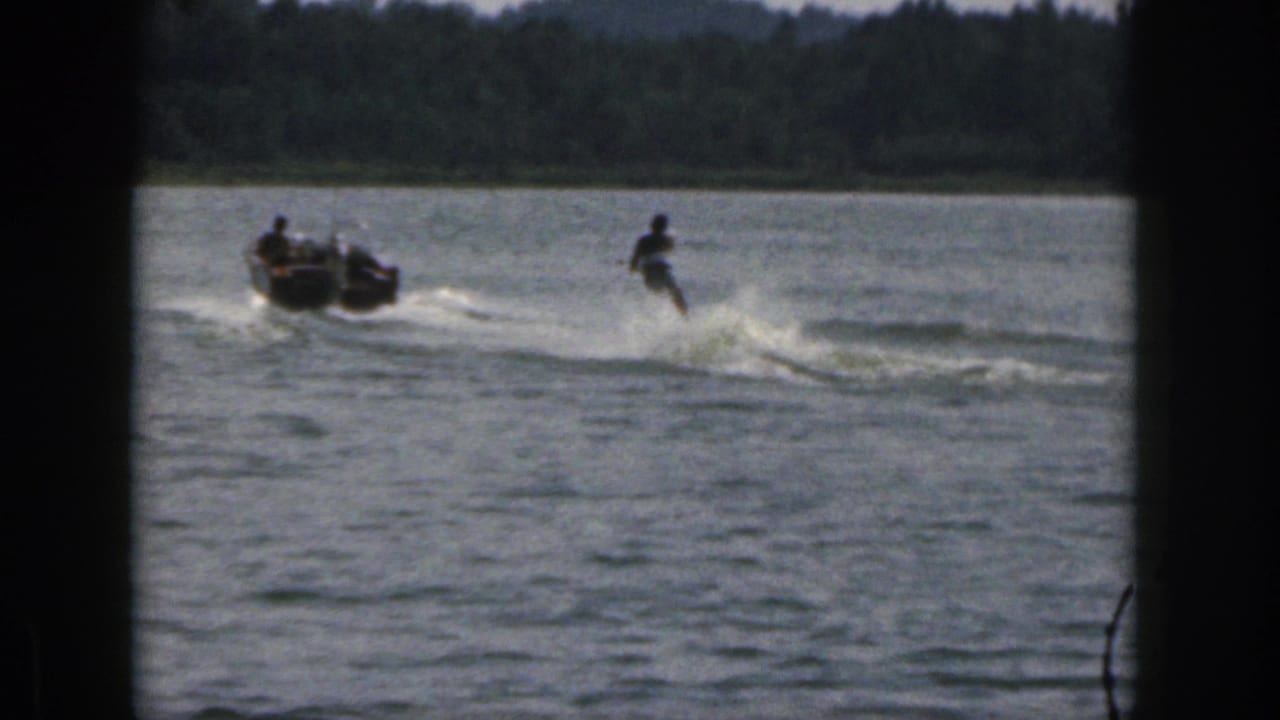 1961 Michigan home-movie frame showing a water-skier being towed by a small motorboat on a wooded lake.