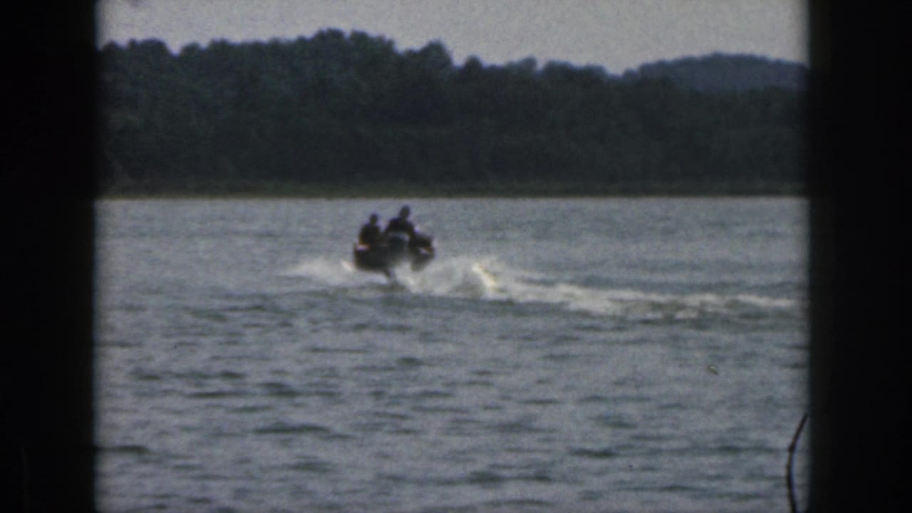 Small motorboat moves across a lake, leaving a wake near a distant tree line.