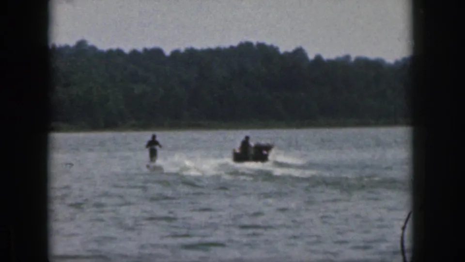1961 Michigan home-movie frame showing the skier and motorboat separated across the lake as the tow line stretches between them.