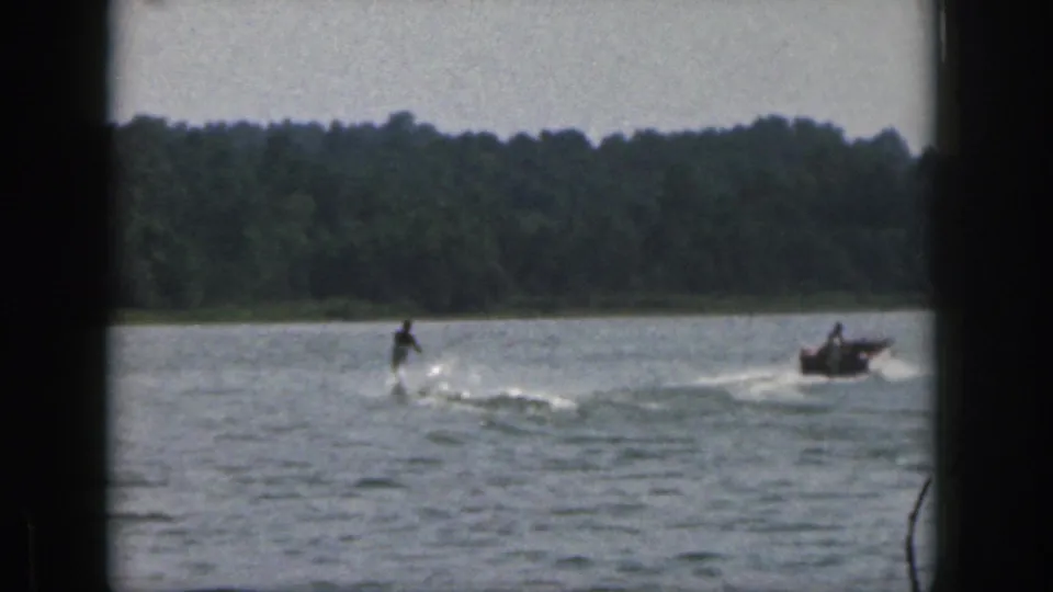 1961 Michigan lake frame with a distant skier trailing a motorboat against a wooded shoreline.