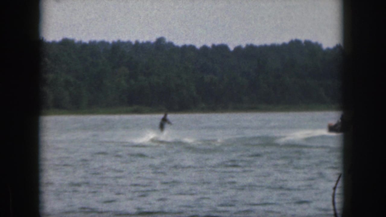 1961 Michigan home-movie frame showing a distant water skier moving across a lake with a forested shoreline in the background.