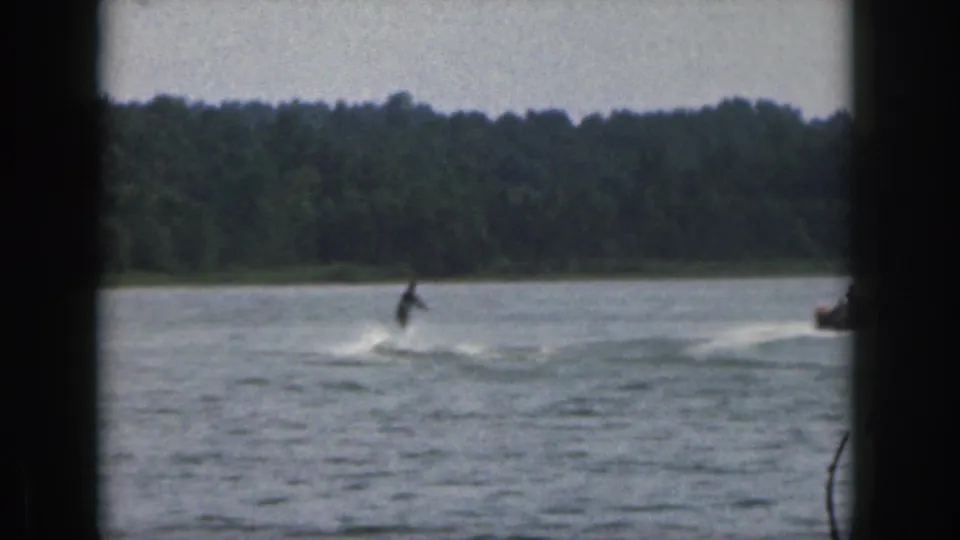 1961 Michigan home-movie frame showing a distant water skier moving across a lake with a forested shoreline in the background.