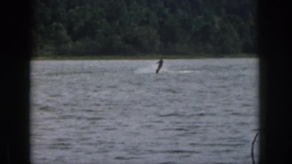 1961 Michigan home-movie frame showing a distant water skier leaning across the lake, with wake and wooded shore visible.